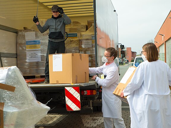 Ein Lkw hält; ein Zusteller übergibt vom Laderaum Kartons. Zwei Menschen in weißen Kitteln und Masken nehmen eine große Kiste entgegen – Lieferung von medizinischem Material.