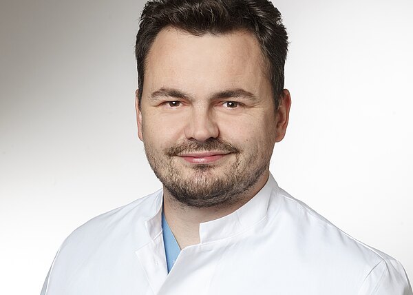 Porträt eines freundlich lächelnden Mannes in weißem Arztkittel vor neutralem Hintergrund; Kopf-und-Schulter-Aufnahme, vermittelt einen professionellen, medizinischen Eindruck.