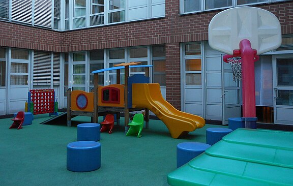 Kinderspielbereich in einem Innenhof: buntes Klettergerüst mit Rutsche, kleine Plastikstühle und Polsterpodeste auf weichem Boden. Rechts ein Basketballkorb, daneben große Schaumstoffmatten. Umgebung wirkt sicher und für Vorschulkinder geeignet.