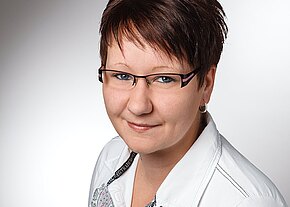 Porträtfoto: lächelnde Frau mit kurzer brauner Frisur und rechteckiger Brille, trägt eine weiße Bluse. Halbprofil vor neutralem, hellgrauem Hintergrund.