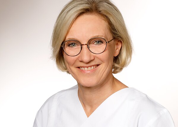 Porträt einer lächelnden Frau mittleren Alters mit kurzem blondem Haar und runder Brille, in weißer medizinischer Kleidung vor hellem, neutralem Hintergrund.