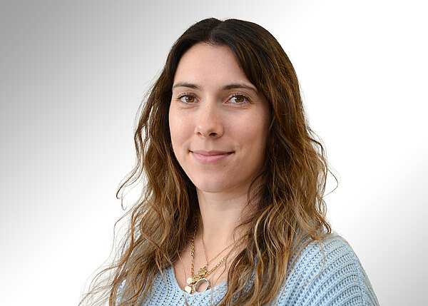 Porträt einer lächelnden Frau mit langen, welligen brünetten Haaren, blickt in die Kamera. Sie trägt einen hellblauen Strickpullover und mehrere goldene Ketten. Heller, neutraler Hintergrund.