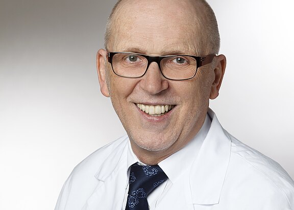 Porträt eines lächelnden, glatzköpfigen Mannes mit Brille in weißem Arztkittel und dunkler Krawatte vor neutralem Hintergrund.