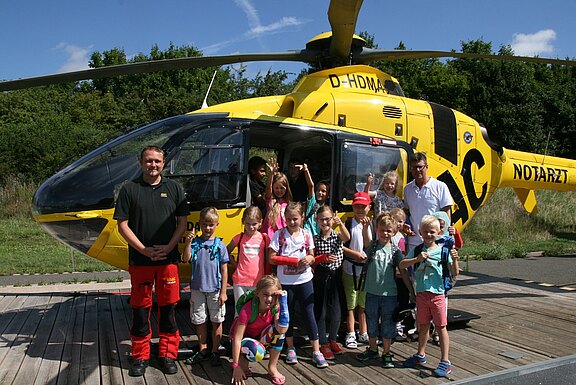 Eine Gruppe fröhlicher Kinder steht mit zwei Erwachsenen vor einem gelben Rettungshubschrauber auf dem Landeplatz. Einige winken, andere zeigen auf den Hubschrauber. Die Szene vermittelt einen Besuch oder eine Führung bei der Luftrettung an einem sonnigen Tag.