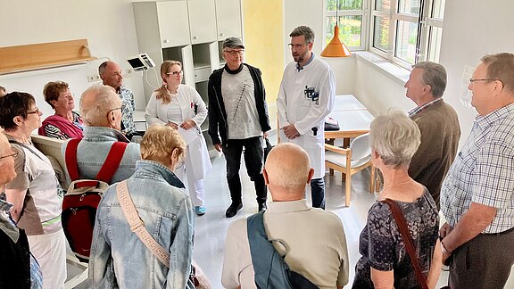 Eine Gruppe älterer Besucher steht in einem Krankenhauszimmer im Kreis. Zwei Mediziner im weißen Kittel sprechen und informieren die Gruppe – eine ruhige Führung bzw. ein Aufklärungsgespräch in der Klinik.