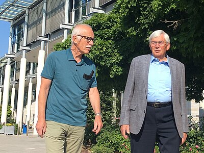 Zwei ältere Männer gehen nebeneinander im Freien vor einem modernen Gebäude. Der eine trägt ein Poloshirt und helle Hose, der andere Sakko und Hemd. Sie wirken im Gespräch. Sonniger Tag, Bäume spenden Schatten.