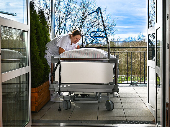 Pflegekraft in weißer Kleidung richtet ein Krankenbett auf einer überdachten Terrasse im Freien aus. Die Türen stehen offen, Tageslicht und Bäume im Hintergrund. Szene vermittelt fürsorgliche Pflege mit Zugang zu frischer Luft.