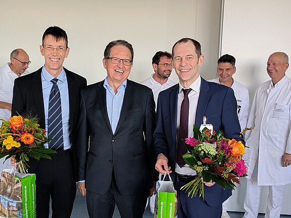 Drei Männer in Anzügen stehen im Vordergrund, jeder mit einem Strauß Blumen in der Hand. Sie lächeln freundlich. Im Hintergrund sind mehrere Personen in weißen Kitteln sichtbar, die ebenfalls lächeln. Die Szene vermittelt eine festliche und herzliche Atmosphäre.
