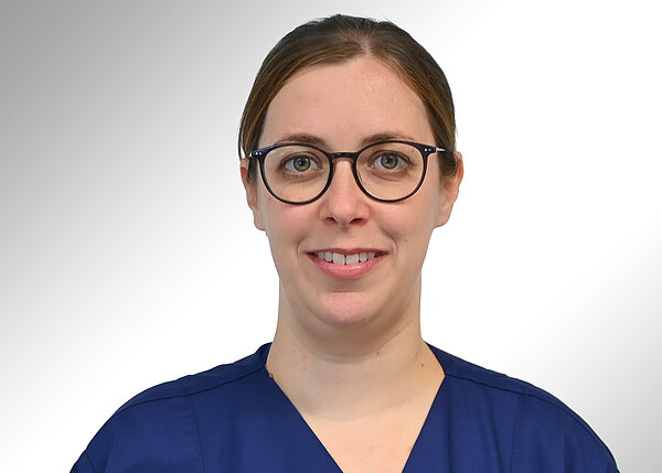 Porträtfoto einer Person mit runder schwarzer Brille und zusammengebundenen Haaren, in blauer medizinischer Kleidung, lächelnd vor neutralem, hellgrauem Hintergrund.