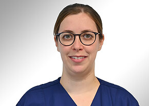 Porträtfoto einer Person mit runder schwarzer Brille und zusammengebundenen Haaren, in blauer medizinischer Kleidung, lächelnd vor neutralem, hellgrauem Hintergrund.