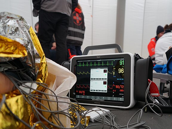 Ein medizinisches Überwachungsgerät zeigt Vitalzeichen wie Herzfrequenz und Blutdruck an. Im Hintergrund sind Rettungskräfte sichtbar, die sich um einen Patienten kümmern. Kabel und ein reflektierendes Material liegen in der Nähe des Geräts.