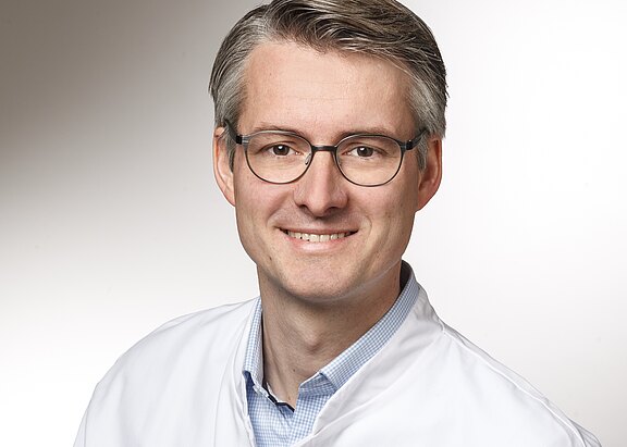 Porträt eines lächelnden Mannes mit Brille in weißem Kittel, blickt in die Kamera vor neutralem Hintergrund.