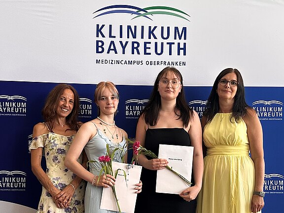 Vier Frauen stehen vor einer Klinikum-Bayreuth-Wand. Zwei junge Frauen in der Mitte halten Urkunden und Blumen, lächeln in die Kamera. Feierliche Stimmung, vermutlich Abschluss- oder Auszeichnungsfeier im medizinischen Umfeld.