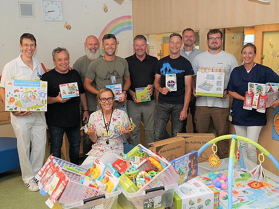 Gruppe von Erwachsenen in einem Kinderkrankenhausraum, die Spielzeugspenden präsentieren; vorne Körbe und Kartons mit bunten Spielen und Babyartikeln; eine Pflegekraft und andere Helfende lächeln, während sie die gespendeten Spielsachen zeigen.