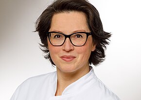 Sumy Büchner Portrait einer Frau mit kurzen, dunklen Haaren und Brille. Sie trägt eine weiße Kitteljacke und lächelt freundlich. Der Hintergrund ist hell und neutral, was den Fokus auf ihr Gesicht lenkt.