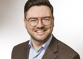 Andreas Thanner Porträt eines lächelnden Mannes mit runder Brille und kurzem Bart. Er trägt ein hellblaues Hemd und ein braunes Sakko vor neutral hellem Hintergrund.