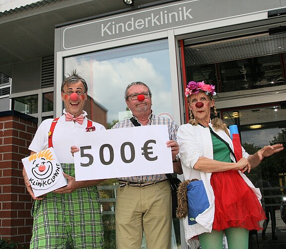 Vor dem Eingang einer Kinderklinik präsentieren zwei Klinikclowns und ein Mann mit roter Nase ein Schild mit „500€“. Die fröhliche Szene deutet auf eine Spende oder Unterstützung für die Kinderstation hin.