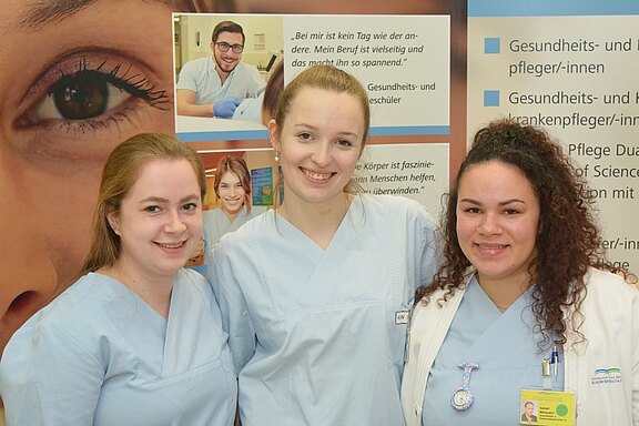 Drei Frauen in hellblauer Berufsbekleidung stehen zusammen und lächeln. Sie repräsentieren den Gesundheits- und Pflegebereich. Im Hintergrund sind Informationsmaterialien zu den Berufen in der Gesundheitsversorgung sichtbar.