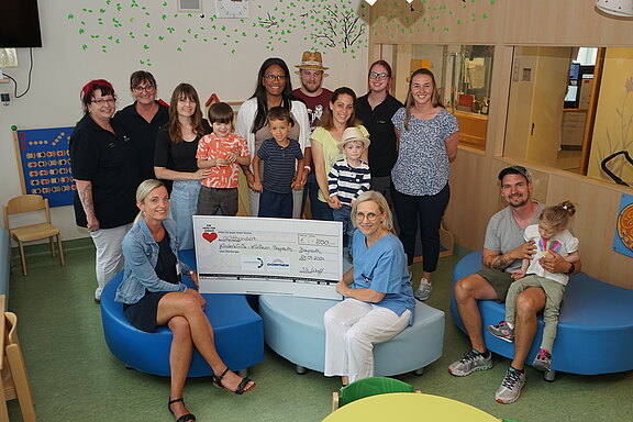 Gruppenfoto in einem Kinderbereich: Erwachsene und Kinder stehen und sitzen zusammen. Im Vordergrund präsentieren zwei Frauen einen großen Spendenscheck. Die Szene zeigt die Übergabe einer Spende an eine Einrichtung für Kinder.