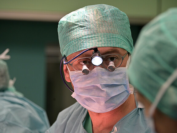 Chirurg im OP trägt grüne OP-Kleidung, Haube und Mundschutz. Er blickt konzentriert, nutzt Lupenbrille mit Stirnlampe. Im Hintergrund unscharf weiteres medizinisches Personal. Szene während einer Operation.