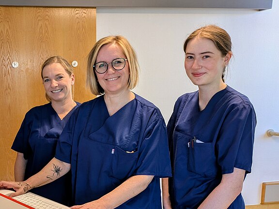 Drei Personen in blauen Pflegeuniformen stehen lächelnd an einem Computerarbeitsplatz in einer Klinik und blicken zur Kamera; auf dem Tisch liegen medizinische Utensilien.