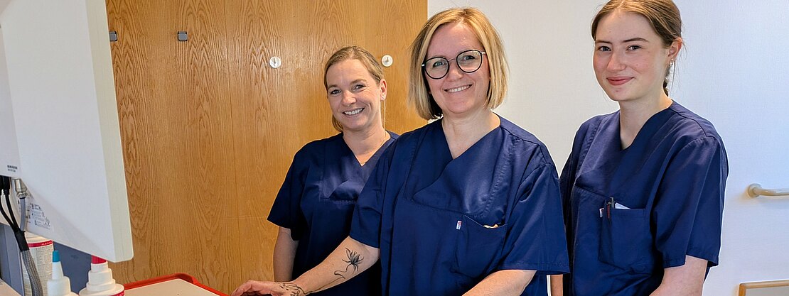 Drei Personen in blauen Pflegeuniformen stehen lächelnd an einem Computerarbeitsplatz in einer Klinik und blicken zur Kamera; auf dem Tisch liegen medizinische Utensilien.