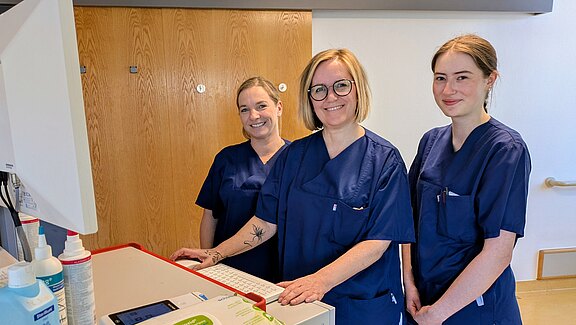 Drei Personen in blauen Pflegeuniformen stehen lächelnd an einem Computerarbeitsplatz in einer Klinik und blicken zur Kamera; auf dem Tisch liegen medizinische Utensilien.