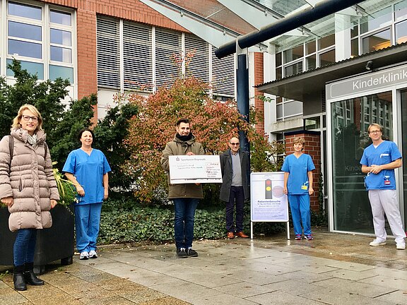 Vor dem Eingang einer Kinderklinik stehen mehrere Personen, darunter Pflegekräfte in blauen Kitteln. Eine Person in der Mitte hält einen großen symbolischen Scheck. Es wirkt wie eine Spendenübergabe zugunsten der Klinik. Regenwetter, Abstand zwischen den Beteiligten.