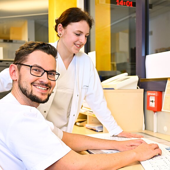 Zwei Personen in Krankenhausuniformen stehen an einem Schreibtisch. Eine Person lächelt in die Kamera, während die andere auf ein Dokument schaut. Der Schreibtisch ist mit Unterlagen und Computerzubehör ausgestattet. Es ist ein freundlicher, arbeitsreicher Moment im Klinikumfeld.