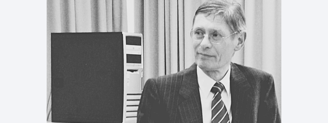 Schwarz-weiß-Foto: Ein älterer Mann im Anzug mit gestreifter Krawatte sitzt in einem Innenraum und blickt nach rechts. Links daneben steht ein Desktop-Computergehäuse. Neutrale, sachliche Szene.