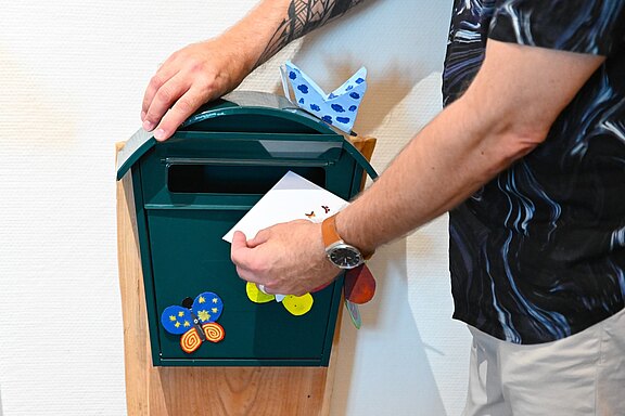 Eine Person hält den geöffneten Deckel eines grünen Briefkastens und wirft einen weißen Umschlag ein. Der Kasten ist mit bunten, kinderhaft gestalteten Schmetterlingen dekoriert.