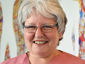 Porträt einer lächelnden Frau mit grauem Kurzhaar und Brille. Sie trägt ein rosafarbenes Kasack-Oberteil mit einem Möwen-Anstecker. Unscharfer, bunter Hintergrund; Fokus auf ihr freundliches Gesicht.