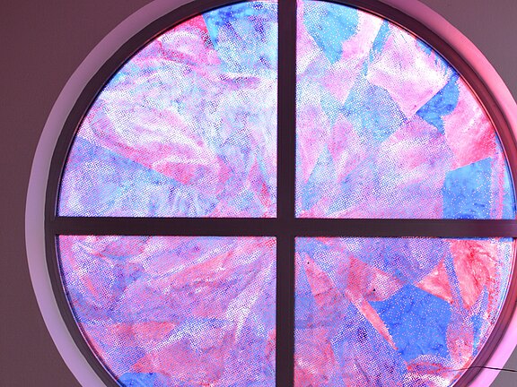 Rundes Fenster mit Kreuzsprossen; die vier Segmente zeigen leuchtende, wolkige Muster in Rosa, Pink und Blau, wie gefärbtes Glas, von Licht durchschienen.
