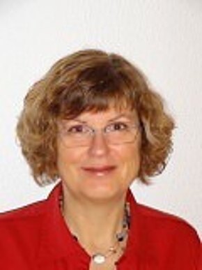 Porträtfoto einer lächelnden erwachsenen Frau mit kurzen, lockigen, hellbraunen Haaren und Brille. Sie trägt eine rote Bluse und eine Halskette. Neutraler, heller Hintergrund; Kopf und Schultern frontal im Bild.