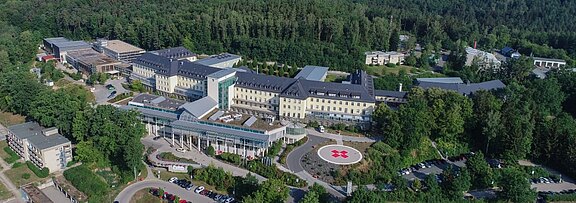 Luftaufnahme eines modernen, mehrstöckigen Gebäudes inmitten von Bäumen. Vor dem Gebäude befindet sich ein großer Platz mit einem roten Kreuz-Symbol. Um das Gebäude sind mehrere Nebenstrukturen und Parkplätze zu sehen, die insgesamt eine einladende und ruhige Einrichtung darstellen.