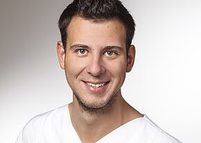 Porträtfoto: junger Mann mit kurzen dunklen Haaren lächelt in die Kamera. Er trägt ein weißes V‑Oberteil vor neutralem, hellem Hintergrund. Fokus auf freundlichem Gesichtsausdruck.