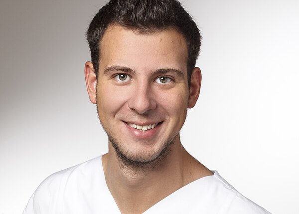 Porträtfoto: junger Mann mit kurzen dunklen Haaren lächelt in die Kamera. Er trägt ein weißes V‑Oberteil vor neutralem, hellem Hintergrund. Fokus auf freundlichem Gesichtsausdruck.