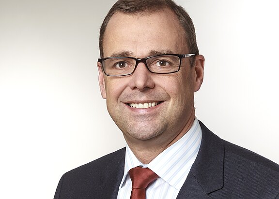 Professionelles Porträt: lächelnder Mann mit Brille, kurzen braunen Haaren, glatt rasiert. Er trägt dunklen Anzug, weißes Hemd und rote Krawatte vor hellem, neutralem Hintergrund.