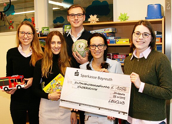 Fünf junge Erwachsene in einem Spielzimmer überreichen einen großen Spendenscheck. Betrag 1.056 Euro, ausgestellt von Sparkasse Bayreuth, Spender: Leo Club, Empfänger: Kinderklinik, Datum 25.01.2019. Einige halten Spielzeug wie Feuerwehrauto und Ball.