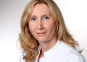 Porträtaufnahme einer blondhaarigen Frau im weißen Kittel, neutraler Blick in die Kamera vor hellem Hintergrund.