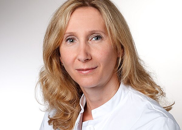 Porträtaufnahme einer blondhaarigen Frau im weißen Kittel, neutraler Blick in die Kamera vor hellem Hintergrund.