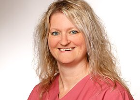 Porträtfoto: Lächelnde Frau mit schulterlangem, welligem, blondem Haar, trägt ein rosa V-Ausschnitt-Oberteil. Sie blickt direkt in die Kamera vor neutralem, hellem Hintergrund.