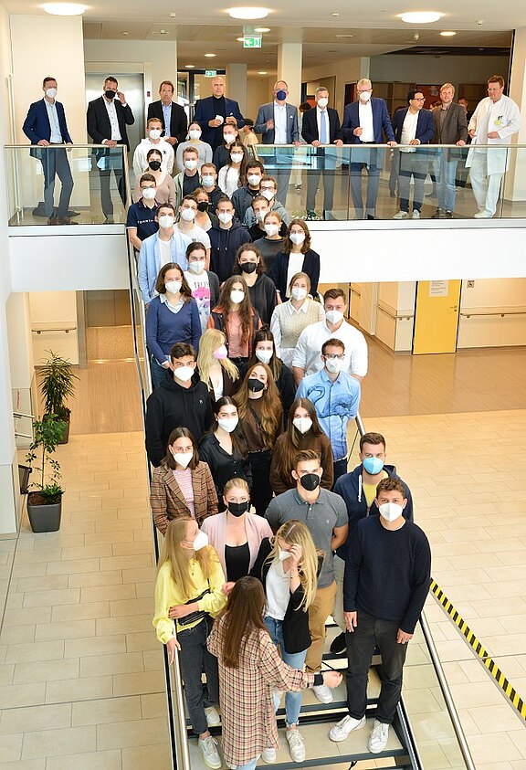 Große Gruppe junger Menschen mit Mund-Nasen-Schutz steht dicht an dicht auf einer Treppe in einem modernen Gebäude und blickt zur Kamera; dahinter weitere Erwachsene auf der Galerie. Gruppenfoto, vermutlich Begrüßung oder Start einer Veranstaltung.
