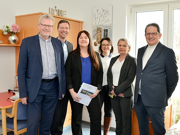 Sechs Personen posieren lächelnd in einem hellen, möblierten Zimmer; eine Person in der Mitte hält eine Broschüre.