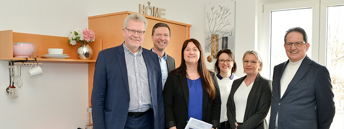 Sechs Personen posieren lächelnd in einem hellen, möblierten Zimmer; eine Person in der Mitte hält eine Broschüre.