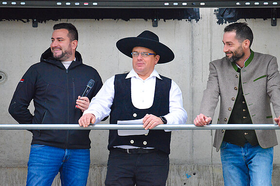 Drei Männer stehen an einem Geländer. Der Mann in der Mitte trägt eine traditionelle Kleidung mit einem schwarzen Hut und hält ein Blatt Papier. Die anderen beiden Männer lächeln, einer hat ein Mikrofon in der Hand, während der dritte eine graue, traditionelle Jacke trägt.