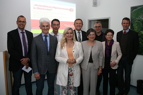 Neun Erwachsene in Businesskleidung posieren in einem Konferenzraum für ein Gruppenfoto. Im Hintergrund zeigt eine Leinwand eine Präsentation mit dem Titel „Weiterbildung Allgemeinmedizin in Oberfranken“. Die Personen stehen dicht zusammen und lächeln, als ob sie eine Fortbildungsveranstaltung abschließen.