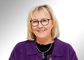 Sabine Jungwirth Porträt einer lächelnden Frau mit Brille und schulterlangem blondem Haar. Sie trägt eine lila Jacke, ein schwarzes Oberteil und eine silberne Halskette. Heller, neutraler Hintergrund.