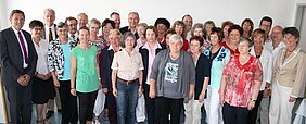 Eine Gruppe von etwa 40 Menschen steht in einem Raum. Die Mehrheit sind ältere Frauen, die in verschiedenen Farben gekleidet sind. Einige Männer sind ebenfalls anwesend. Alle schauen in Richtung der Kamera und lächeln. Der Hintergrund ist hell und neutral.