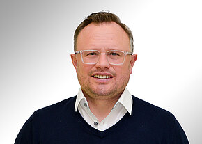 Florian Bär Porträt eines lächelnden Mannes mit kurz geschnittenen Haaren, heller Kunststoffbrille, weißem Hemd und dunkelblauem Pullover, frontal vor neutralem, hellem Hintergrund.